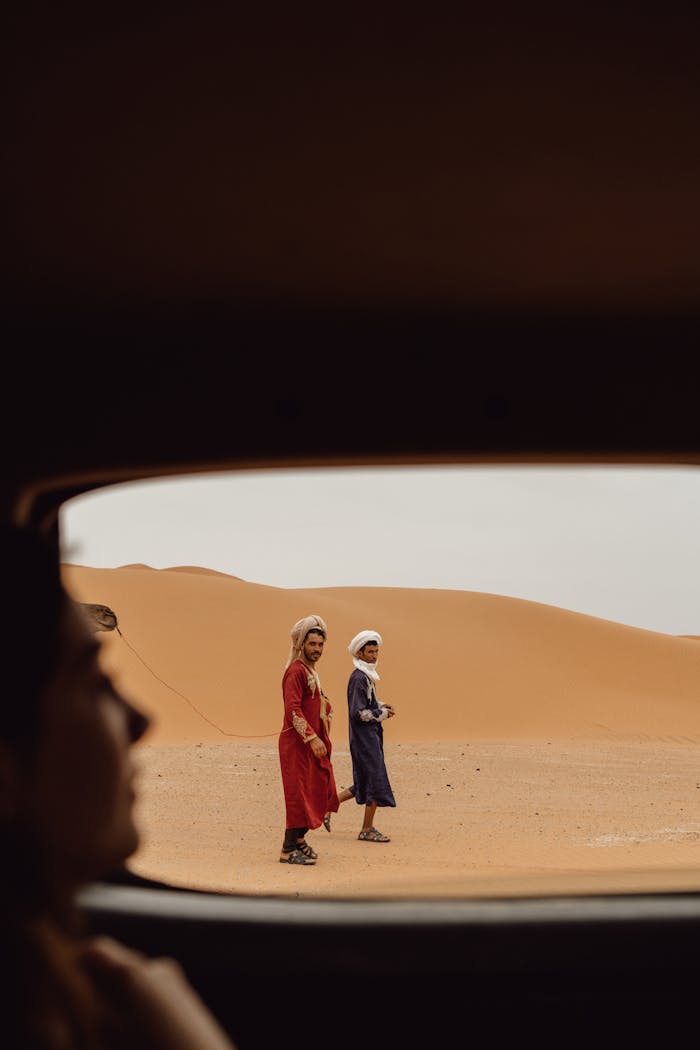 Two men in traditional attire seen through a car window in Merzouga Desert.